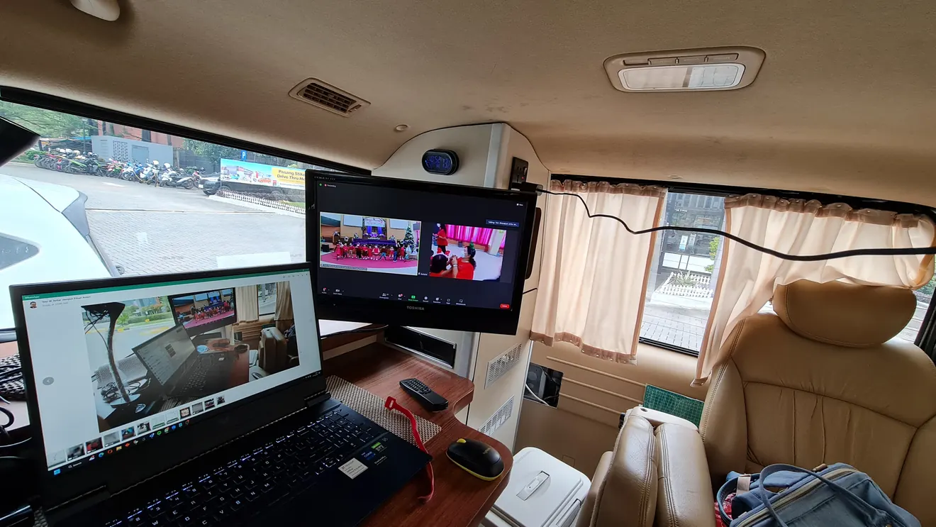 Bekerja dari dalam H1 di area parkir: laptop di meja kayu, monitor tambahan menampilkan meeting Zoom, dan jendela memperlihatkan halaman parkir di luar